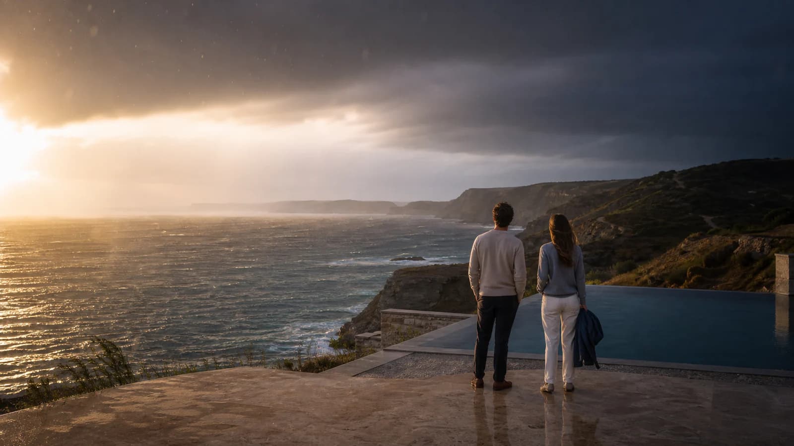 Two travellers watching sunlight break through a coastal storm beside a cliffside infinity pool